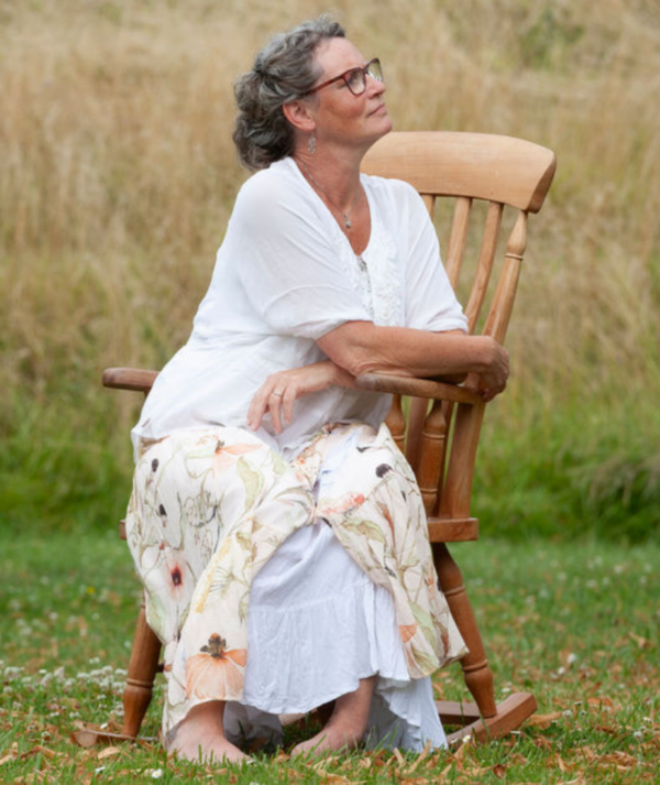 Vrouw in witte kleding zit buiten op een houten schommelstoel in het gras.