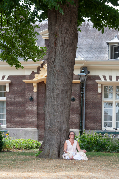 Vrouw zit aan de voet van een grote boom voor een gebouw.